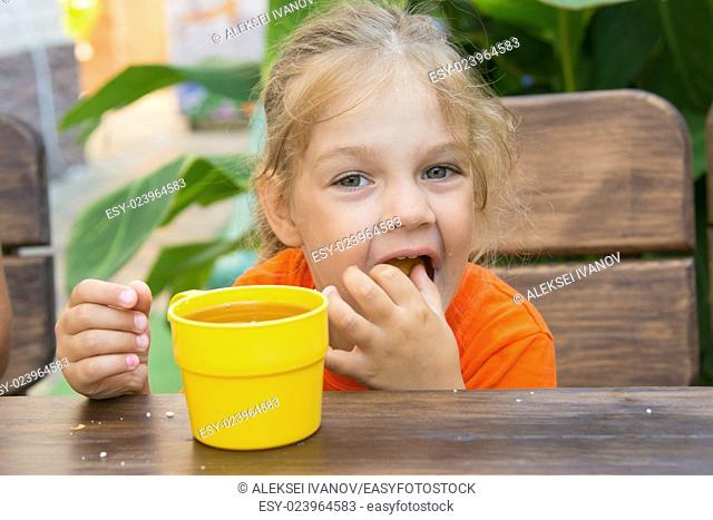 Four-year girl sitting at a wooden table on the porch, drinking a drink from the cup and eating a bun