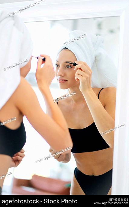Woman in lingerie and a head towel in front of a mirror applies makeup