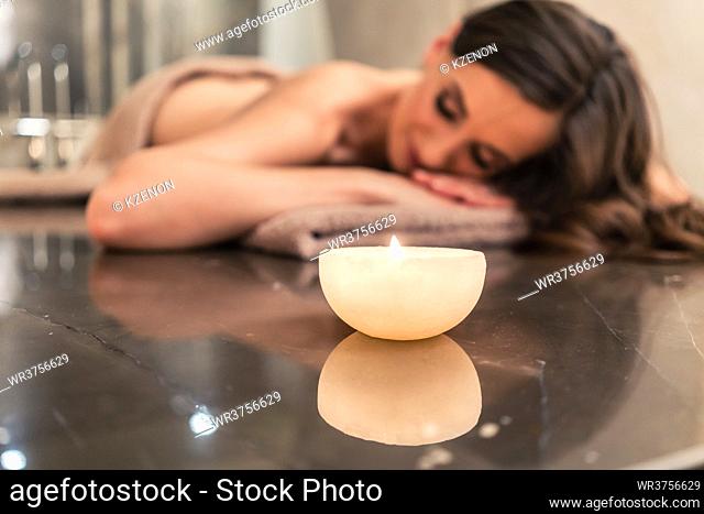 Scented round candle next to young woman on marble massage table during aromatherapy at luxury spa and wellness center