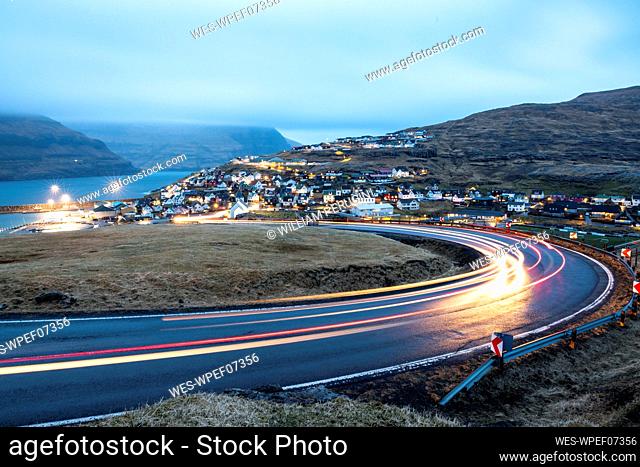LIght trails on road with Eidi town in background at dusk, Faroe Islands