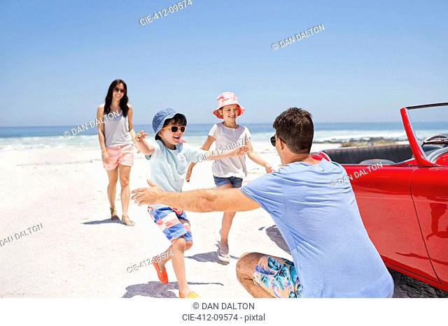 Father reaching to hug children on beach next to convertible