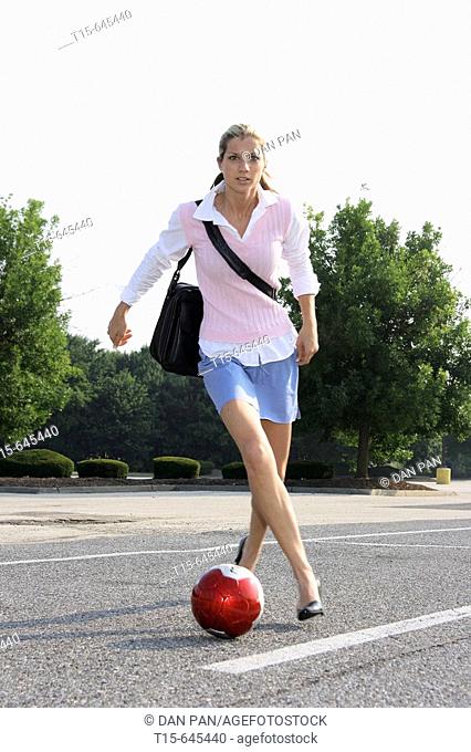 Girl in her 20's in work clothes carrying a laptop playing soccer in parking lot on her way to work