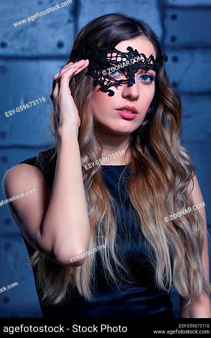 Young woman in black lace mask and black dress. Blonde female with long hair and hands near face. Female on smoke and metal wall background