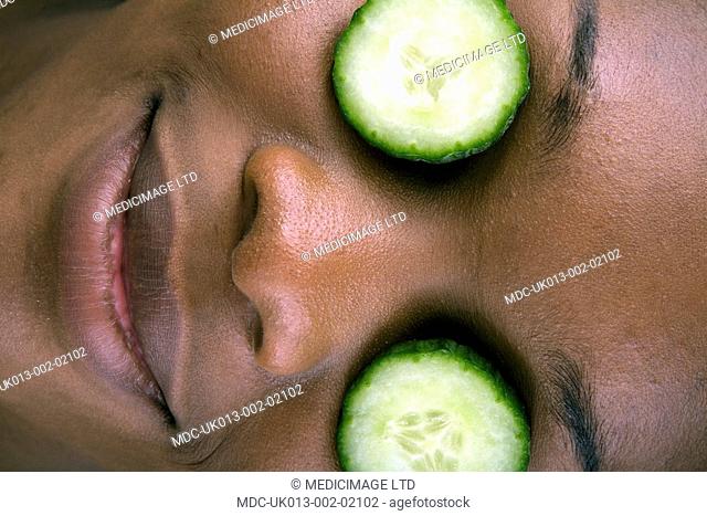 A young black girl using slices of cucumber as a cosmetic because it is a trong source of vitamin C, vitamin K, potassium, vitamin A, vitamin B6, thiamin