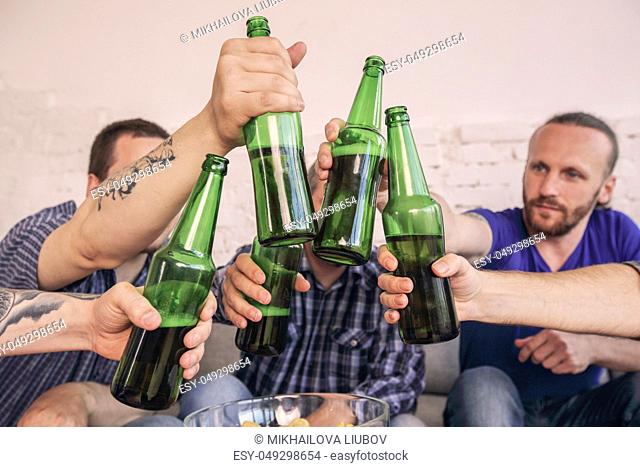 Group of men clinking beer, eating pizza, talking and smiling while resting at home on couch behind TV