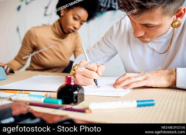 Hipster man writing with fountain pen on paper while sitting by woman in studio