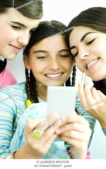 Three young female friends looking at selves in hand mirror, one holding lipgloss