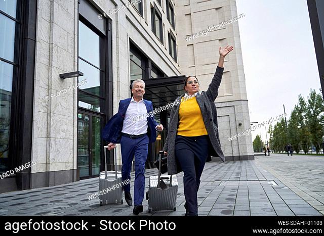 Business colleagues running with wheeled luggage on footpath while waving in city
