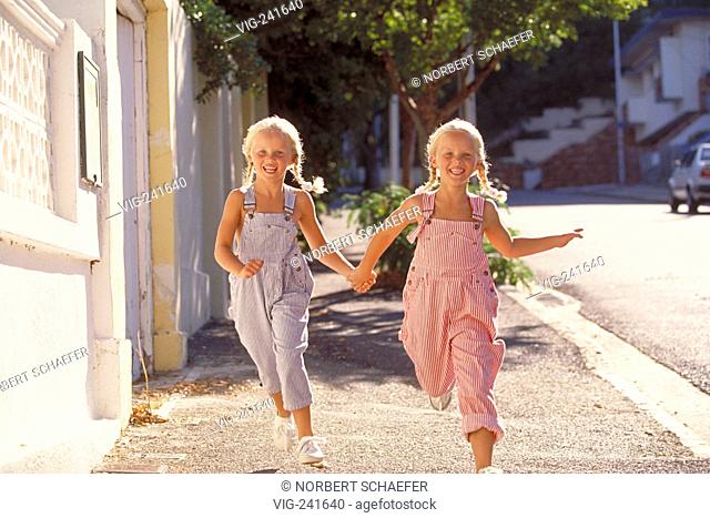 portrait, full-figure, two 5-year-old twin-girls with blond plaits wearing blue and red-white striped trousers with braces run laughing hand-in-hand along the...