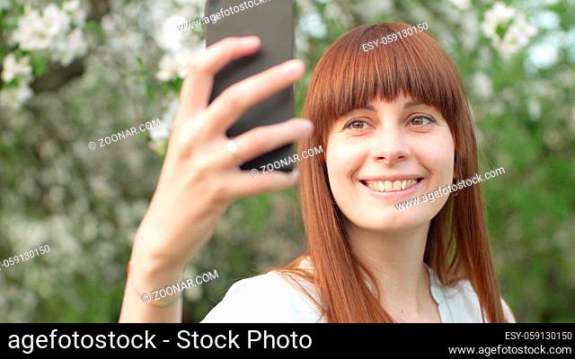 A girl makes selfie in the garden. An attractive red-haired woman smiles making selfi using a mobile phone in a cherry orchard