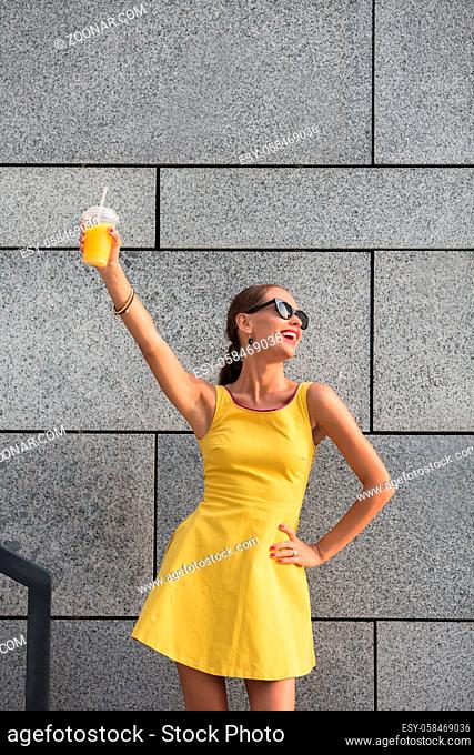 Stylish woman in sunglasses drinking orange cockrail on the street. Happy woman in little yellow dress posing with her hand on hip