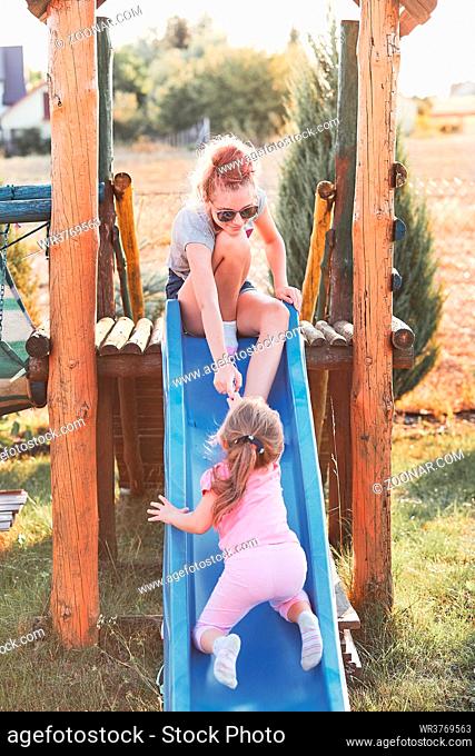 Teenage girl playing with her younger sister in a home playground in a backyard. Happy smiling sisters having fun on a slide together on summer day