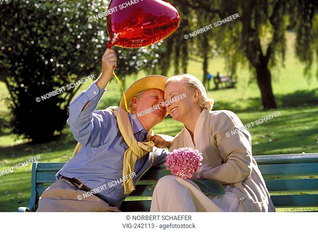 park-scene, outdoor, senior-couple wearing bright dresses and strawhat sits with a heart shaped balloon and lilac flowers on a parkbench  - GERMANY, 23/08/2004