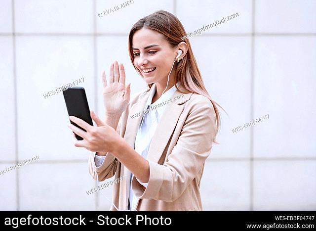 Businesswoman waving hand during video call through smart phone in front of wall