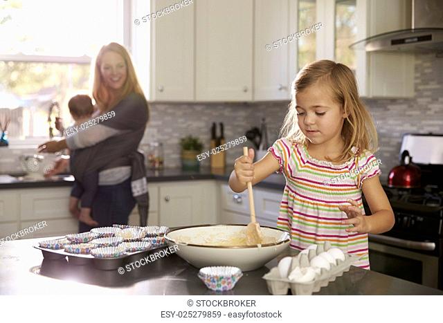 Young girl prepares cake mix, mum and baby in the background