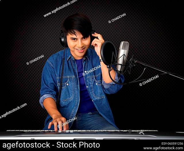 Young asian man with headphones playing an electric keyboard in front of black soundproofing walls. Musicians producing music in professional recording studio