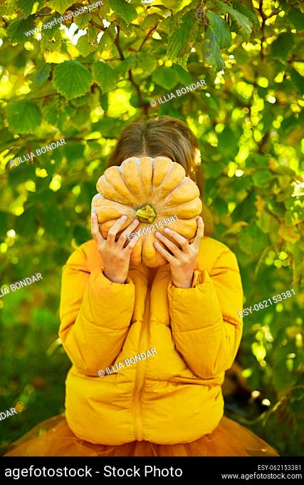 Happy child girl covers the face with pumpkin outdoors in halloween, An orange pumpkin in the hands of a girl in the autumn park