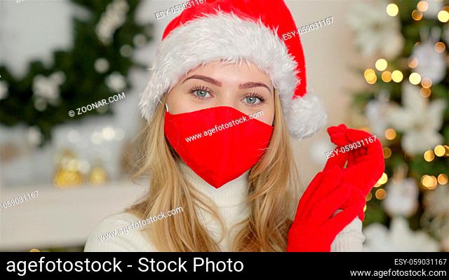 Portrait of a beautiful woman in christmas hat while standing over cristmas tree background. The woman wears the protective mask from the virus