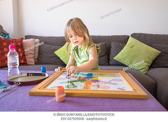 four years old blonde child with green sleeveless shirt sitting on brown sofa, playing parcheesi or parchisi or parchis, on table