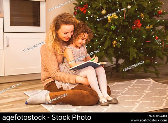 Lovely mother and daughter near Christmas tree. Young mother holding her daughter on her knees, helping to draw a greeting card for Christmas