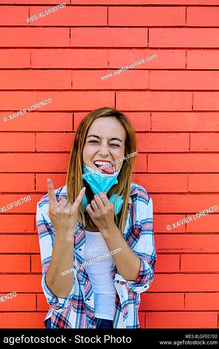 Woman sticking out tongue while horn sign gesture standing against wall