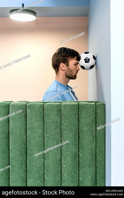 Male entrepreneur pressing soccer ball with head on wall at workplace