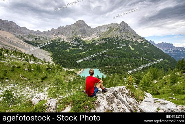Young man, hiker sitting on rocks and looking at turquoise green Sorapis lake and mountain landscape, Dolomites, Belluno, Italy, Europe