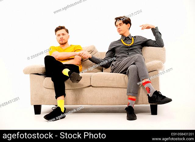 Two cute guys make a bet hand in hand sitting on a white sofa. Group of friends are sitting on a soft couch and communicates isolated on white background