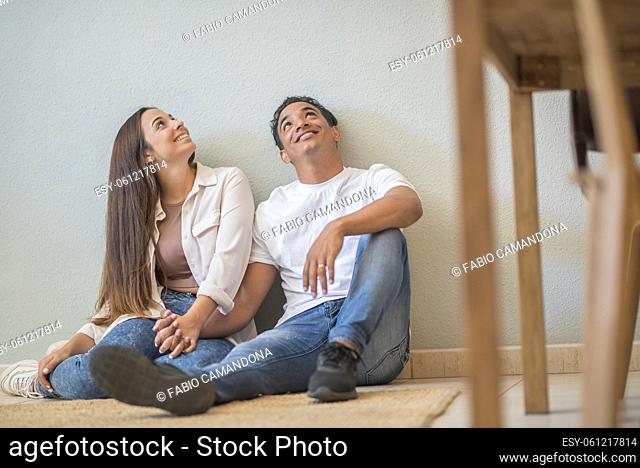 Young couple at home sitting on the floor looking up in to the air and smiling - concept of love and relationship wito mortgage and new life together -...