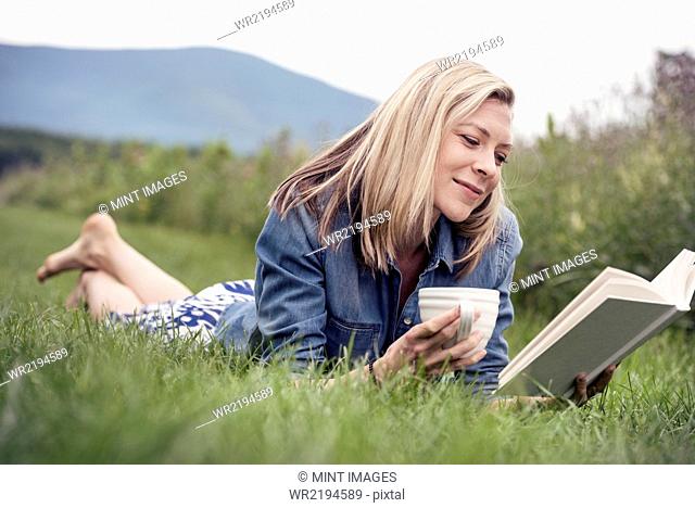 A woman lying on the grass holding a tea cup and reading a book