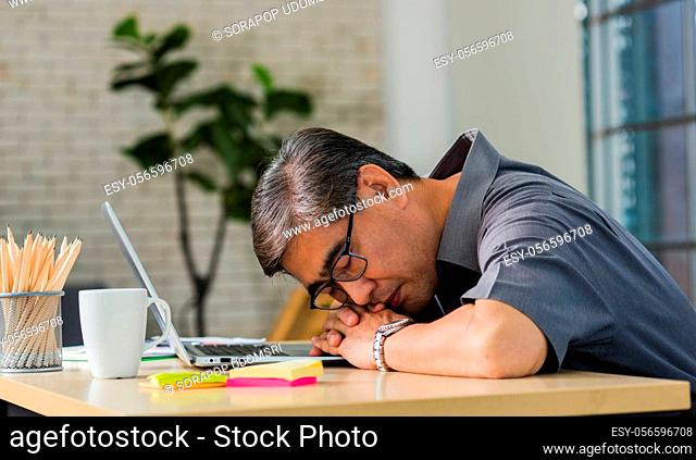 Asian businessman tired overworked he sleeping over a laptop computer on the desk. senior man with eyeglasses lying asleep on table at his working place