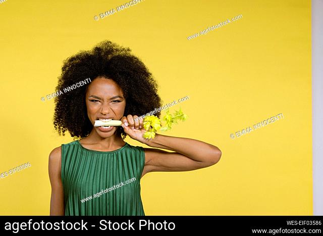 Young woman biting celery against yellow background