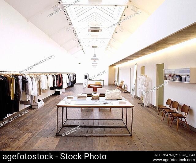 Interior of retail designer clothes shop glass slanted ceiling metal clothes rail lined with clothes Interiors shops retailing metal tables with wooden work...