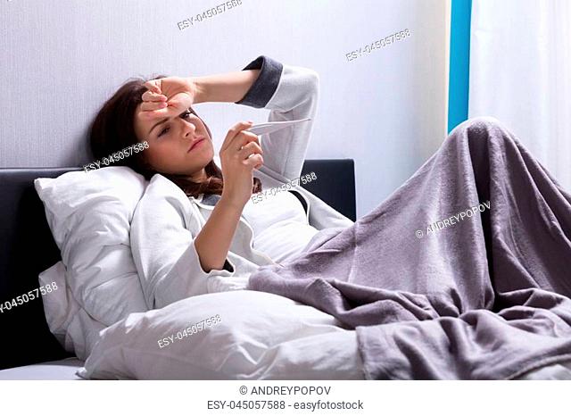 Sick Young Woman Lying On Bed Checking Body Temperature With Thermometer