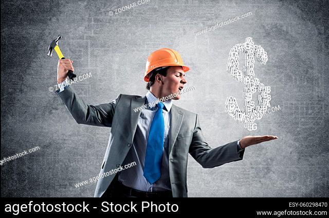 Furious businessman going to crash with hammer dollar symbol. Young handsome man in business suit and safety helmet standing on wall background