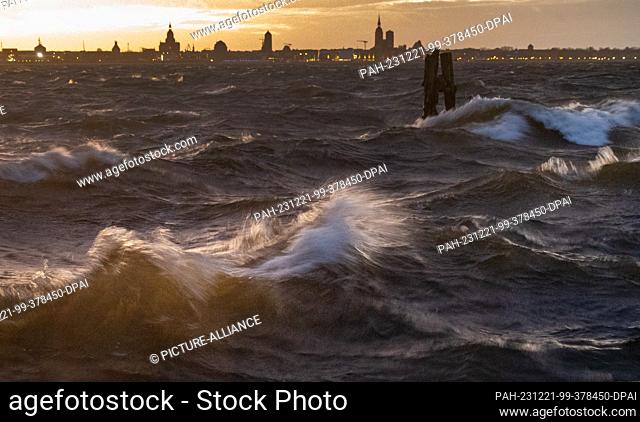 21 December 2023, Mecklenburg-Western Pomerania, Altefähr: Waves break on the beach at Altefähr on the island of Rügen. The silhouette of the Hanseatic city of...