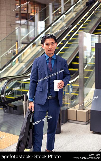 A young businessman in a blue suit on the move in a city downtown area, carrying a briefcase and cup of coffee