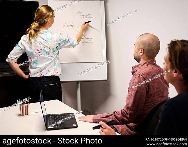 PRODUCTION - 07 July 2021, Berlin: ILLUSTRATION - A woman writes on a flip chart in a meeting room while two men sit at a table
