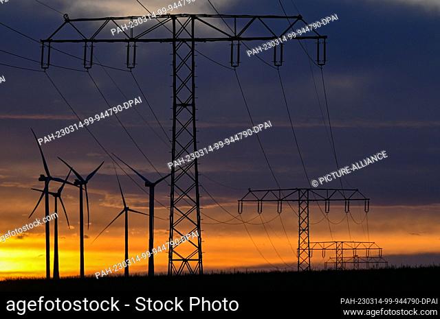 14 March 2023, Brandenburg, Jacobsdorf: Colorful clouds glow in the early morning sunrise over the landscape with several wind turbines and a high-voltage power...