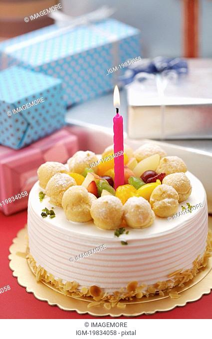 Birthday cake with a lighted candle on top, wrapped presents in the background