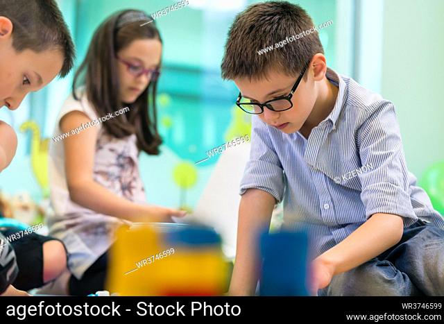 Cute girl wearing eyeglasses while playing with concentration a creative game next to her colleague in the classroom of a modern kindergarten