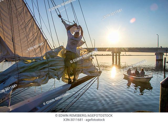 Adult male fixes sail on traditional Skipjack boat as the sun rises above a bridge in the distance