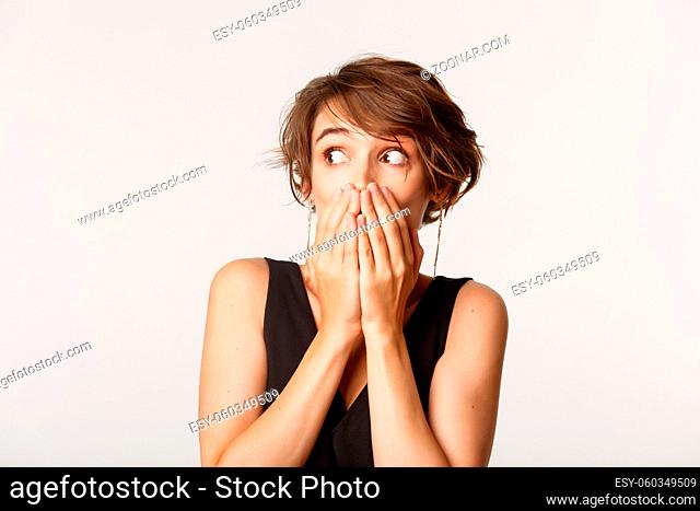 Close-up of shocked gossip girl looking left, cover mouth with hands, standing over white background