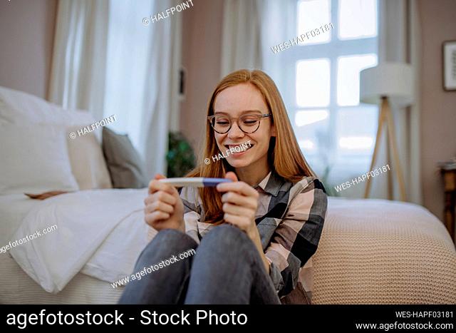 Happy redhead woman with pregnancy testing kit sitting in front of bed at home
