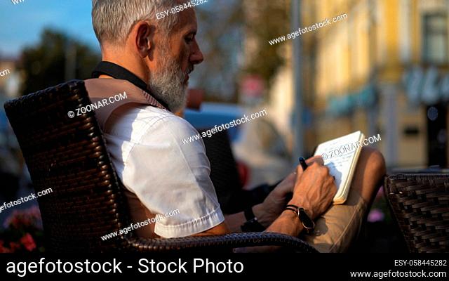 A grey light-skinned bearded man inspiredly writes down ideas in a notebook in a street cafe. Close Up