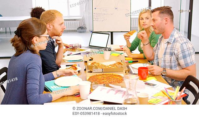 Side view of friendly colleagues relaxing at table in office having ordered pizza and talking lively while resting