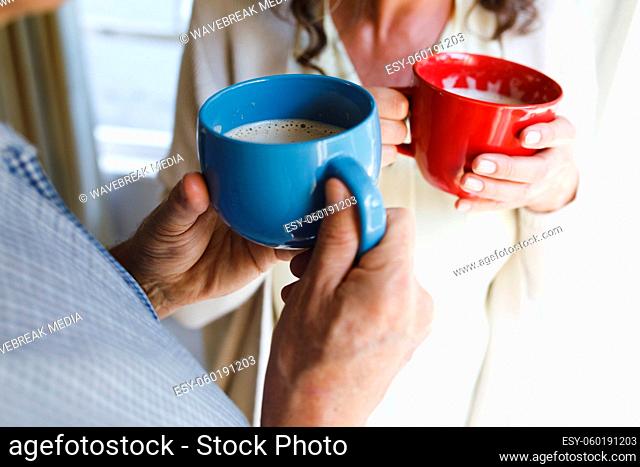 Senior caucasian couple standing next to window, holding cups