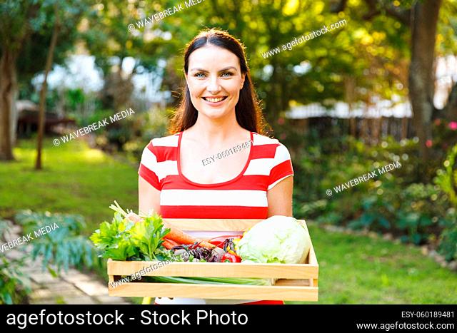 Portrait of smiling caucasian woman standing in garden holding box of fresh organic vegetables