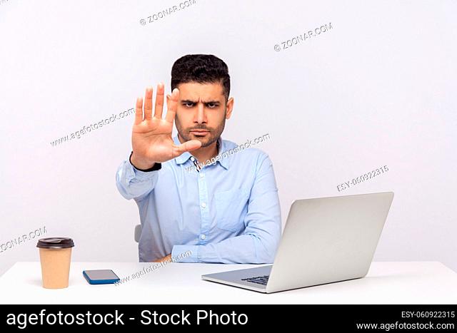 Strict bossy elegant young businessman sitting in office workplace with laptop and coffee, warning with stop gesture, denial, prohibition hand sign