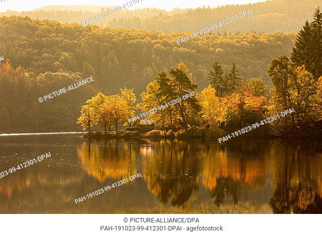 23 October 2019, North Rhine-Westphalia, Simmerath: Autumnally colourful trees and a house are reflected in the Rursee lake near Rurberg